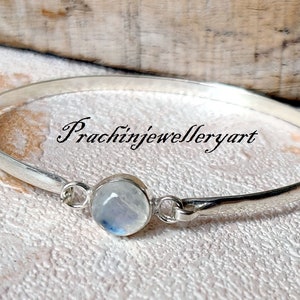 May include: A silver bangle bracelet featuring a round, white moonstone gemstone. The bracelet is open and has a simple design. The text "Prachinjewelleryart" is visible on the bracelet.