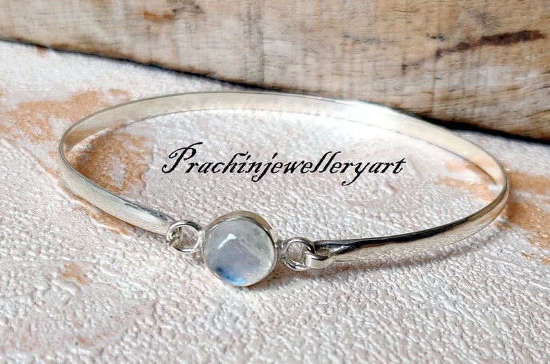 May include: A silver bangle bracelet featuring a round, white moonstone gemstone. The bracelet is open and has a simple design. The text "Prachinjewelleryart" is visible on the bracelet.