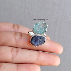 May include: A silver ring with two raw gemstones. The top stone is a light blue aquamarine, and the bottom stone is a dark blue sapphire.