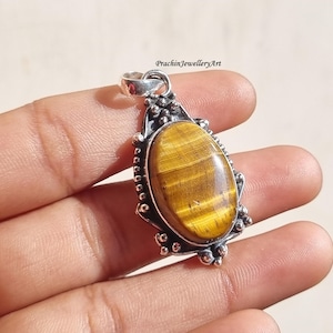 Puede incluir: Un colgante de plata con una piedra de ojo de tigre de forma ovalada. El colgante tiene un marco de plata detallado con un borde de cuentas.