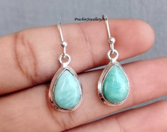 May include: A pair of silver dangle earrings with teardrop-shaped green gemstones. The earrings are on a hand.