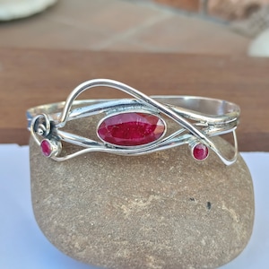 May include: A silver bracelet with three oval-cut, deep red gemstones. The bracelet has an open design with a central large stone and two smaller stones. The metalwork has a flowing, organic style.