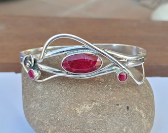 Puede incluir: Una pulsera de plata con tres piedras preciosas rojas oscuras de corte ovalado. La pulsera tiene un diseño abierto con una piedra central grande y dos piedras más pequeñas. La filigrana tiene un estilo fluido y orgánico.
