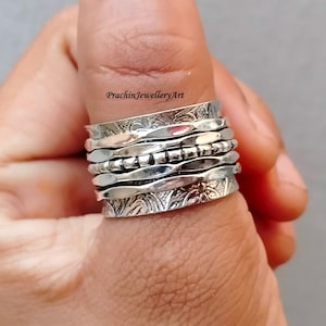 May include: A silver spinner ring with multiple bands, each with a different texture. The top band has a floral design, the middle band has a beaded design, and the bottom band has a hammered finish.