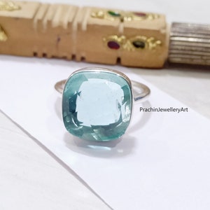 May include: A square-cut, light blue gemstone ring with a silver band. The gemstone is set in a bezel setting, and the ring is displayed on a white surface. The text "PrachinJewelleryArt" is visible on the right side of the ring.