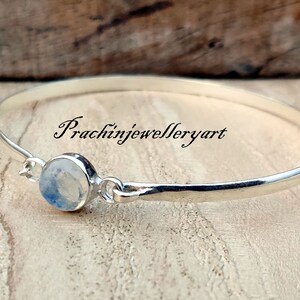 May include: A silver bangle bracelet featuring a moonstone cabochon. The bracelet is open and has a simple design. The text "Prachinjewelleryart" is visible on the bracelet.