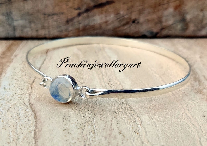 May include: A silver bangle bracelet featuring a moonstone cabochon. The bracelet is open and has a simple design. The text "Prachinjewelleryart" is visible on the bracelet.
