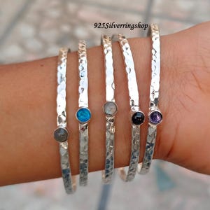 May include: Set of five silver bangles with hammered texture and small gemstone accents. The gemstones are grey, blue, white, black, and purple.