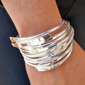Pode incluir: Um conjunto de pulseiras prateadas. As pulseiras são lisas e curvas, com uma pequena abertura nas extremidades. As pulseiras estão empilhadas, criando um efeito em camadas. As joias são exibidas sobre um fundo escuro.