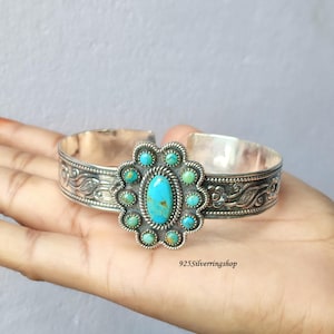 Könnte beinhalten: Ein Silberarmreif mit einem zentralen ovalen Türkisstein, umgeben von einer Gruppe kleinerer runder Türkissteine. Das Band ist mit geprägten Blumenmustern und seilartigen Metalldetails verziert. Der Text lautet 925Silverringshop.