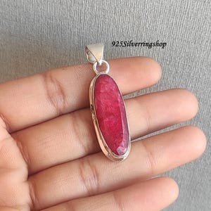 May include: A silver pendant with a large, oval-shaped, faceted ruby gemstone. The ruby is a deep red color and is set in a silver bezel. The pendant has a simple, elegant design. The text "925Silverringshop" is visible.