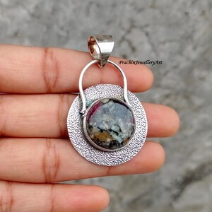 May include: A silver pendant featuring a round, multi-coloured stone set within a textured silver frame. The pendant has a bail for attaching to a chain.
