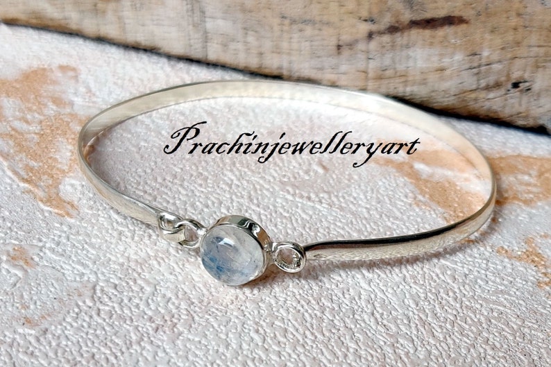 May include: A silver bangle bracelet featuring a round, white moonstone gemstone. The bracelet is open and has a simple design. The text "Prachinjewelleryart" is visible in the image.