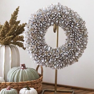 Pode incluir: Uma coroa circular construída a partir de numerosas pequenas vagens de sementes secas brancas e castanho-claras, suspensa num suporte de metal fino de tom dourado com uma base retangular. A coroa apresenta uma aparência densa e texturizada.
