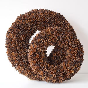 May include: A decorative, abstract sculpture composed of two intertwined circular forms. The sculpture is covered in small, dried, brown leaf-like elements, creating a textured surface. The piece is set against a white background.