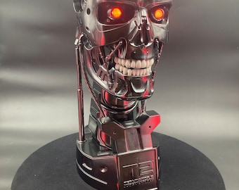 May include: A silver Terminator 2 bust with red glowing eyes. The bust is on a black base with the text "Terminator 2: Judgment Day" on it. There are three small accessories next to the bust.