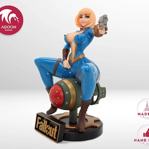 Puede incluir: Figura de un personaje de la serie de videojuegos Fallout. La figura lleva un mono azul y botas marrones, sostiene un arma y está sentada sobre una bomba. La base de la figura tiene la palabra "Fallout".