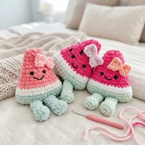 May include: Three handmade crochet watermelon plush toys with smiling faces and bows. The toys are pink, white, and light green, with black seed details. A crochet hook and pink yarn are also visible.