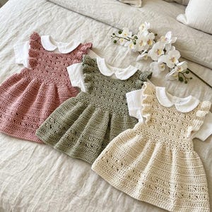 Könnte beinhalten: Drei Häkelkleider in Rosa, Grün und Creme, jedes mit einem weißen Bubikragen und kurzen Ärmeln. Die Kleider sind auf einem Bett mit einer neutralfarbenen Bettdecke drapiert. Ein Zweig weißer Blumen liegt daneben.