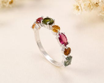 May include: A silver ring featuring a row of seven multi-colored gemstones in various shapes and sizes. The stones include shades of red, green, yellow, and orange, set in a polished silver band that curves to display the stones along the top arc.