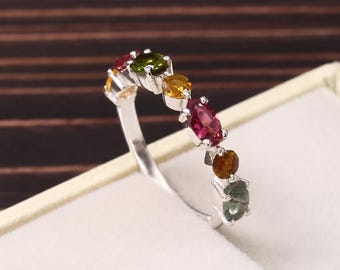 May include: A silver ring featuring a row of seven multi-colored gemstones in various shapes and sizes, including green, yellow, red, and brown hues. The stones are set in individual silver prongs along the top half of the thin, polished band.