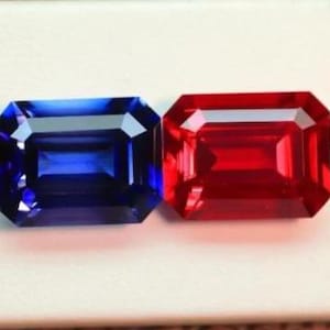 Op de afbeelding: Twee rechthoekige edelstenen, een diepblauwe en een felrode, worden naast elkaar weergegeven. De edelstenen hebben een facetgeslepen slijpvorm, die het licht reflecteert. De achtergrond is een effen wit oppervlak.