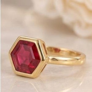 May include: A gold ring featuring a hexagonal-cut deep red gemstone set in a bezel setting. The band is smooth and polished, showcasing a clean, metallic finish that complements the geometric shape of the central stone.