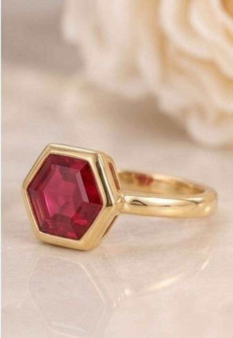 May include: A gold ring featuring a hexagonal-cut deep red gemstone set in a bezel setting. The band is smooth and polished, showcasing a clean, metallic finish that complements the geometric shape of the central stone.