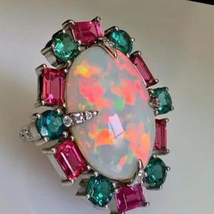 May include: A silver ring with a large, oval opal gemstone displaying internal colours. The opal is encircled by rectangular and round gemstones in pink, teal, and green hues. Small, clear accent stones are also set around the opal.