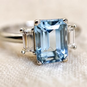 May include: A silver-toned ring featuring a large, rectangular, emerald-cut light blue gemstone at the centre, flanked by two smaller, rectangular, baguette-cut clear stones on either side, all set in a polished metal band with four-prong settings.