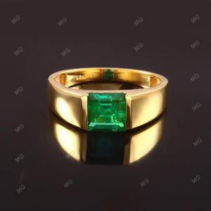 Könnte beinhalten: Ein goldener Ring mit einem großen, quadratisch geschliffenen Smaragd-Edelstein. Der Ring hat ein dickes Band und eine polierte Oberfläche, die das Licht reflektiert. Der Smaragd hat eine leuchtend grüne Farbe und ist in einer Zargenfassung gefasst.