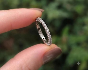 May include: A silver-toned eternity band ring featuring a continuous row of small, round, clear faceted stones set along the entire circumference of the metal band, creating a sparkling and uniform appearance around the circular jewelry piece.