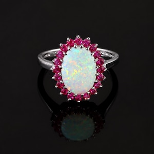 May include: A silver-toned ring featuring a large, oval-shaped white opal centre stone with iridescent flashes of colour. The central opal is surrounded by a halo of small, round, vibrant pinkish-red rubies set in a scalloped metal frame.