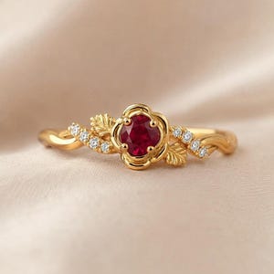 Pode incluir: Um anel de ouro com um rubi vermelho central de corte redondo engastado numa estrutura em forma de flor. O aro é decorado com pequenas pedras claras em ambos os lados e detalhes de folhas esculpidas que se estendem do centro floral.