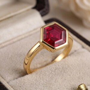 May include: A gold ring featuring a single, faceted, hexagonal-cut deep red gemstone set in a smooth, polished gold bezel. The band is a simple, solid gold shank with a rounded profile, showcasing a clean, geometric design and vibrant colour.