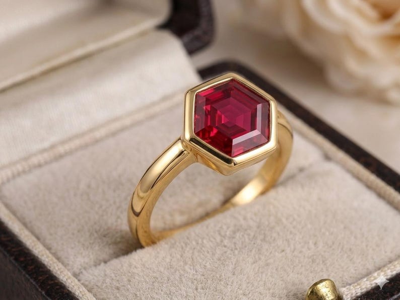 May include: A gold ring featuring a single, faceted, hexagonal-cut deep red gemstone set in a smooth, polished gold bezel. The band is a simple, solid gold shank with a rounded profile, showcasing a clean, geometric design and vibrant colour.