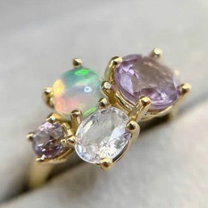 Könnte beinhalten: Ein goldener Ring mit fünf Edelsteinen. Der Ring hat einen großen weißen Opal, zwei kleinere Amethyststeine und zwei kleinere klare Steine.
