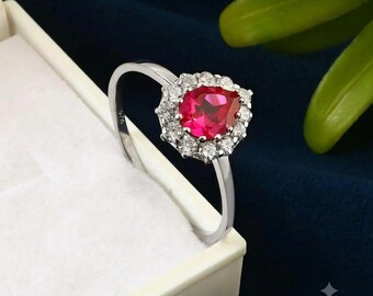 May include: A silver ring featuring a pear-shaped red gemstone at the centre, surrounded by a halo of small clear stones. The band is stamped with 925, indicating sterling silver, and the ring is presented in a white jewellery box.