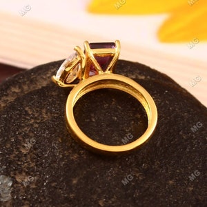 May include: A gold ring with a large purple gemstone and a smaller clear gemstone. The ring is set on a dark brown surface.