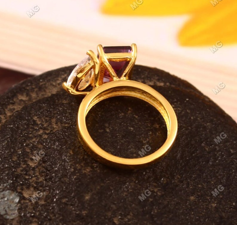 May include: A gold ring with a large purple gemstone and a smaller clear gemstone. The ring is set on a dark brown surface.