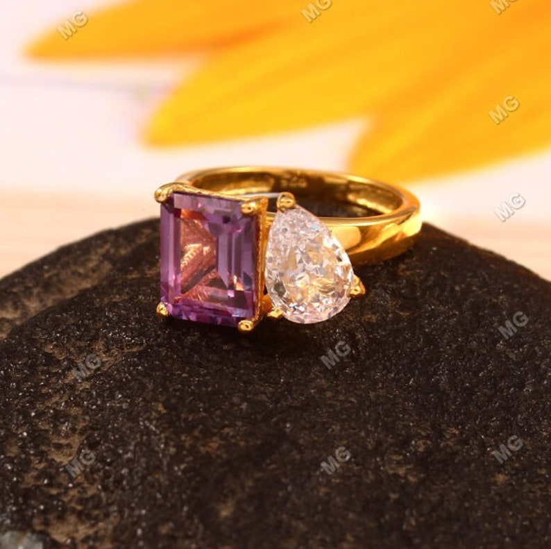 May include: A gold ring with a purple square-shaped gemstone and a clear pear-shaped gemstone.