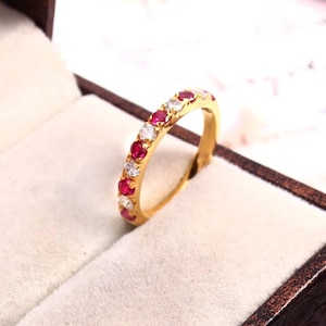 Può includere: Un anello a fascia d'oro con pietre preziose rosse e bianche alternate. L'anello è in una scatola per anelli di velluto bianco.