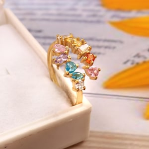 Può includere: Un anello d'oro con un arcobaleno di pietre preziose colorate. L'anello è in una scatola bianca.
