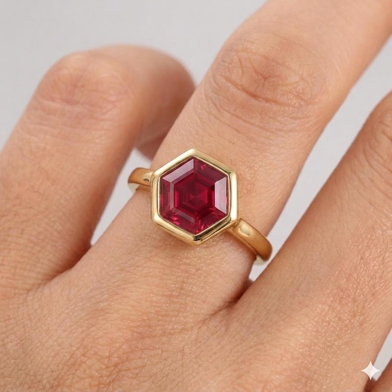 May include: A gold ring featuring a central hexagonal-cut deep red gemstone. The stone is set within a smooth, polished gold bezel setting that follows the hexagonal shape, mounted on a simple, slender, polished gold band.
