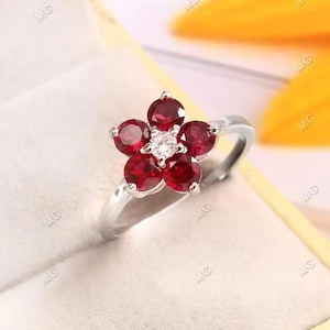 Może przedstawiać: Srebrny pierścionek z motywem kwiatowym. Kwiat ma pięć płatków, każdy osadzony okrągłym, głęboko czerwonym kamieniem szlachetnym. Środek kwiatu to mały, przezroczysty kamień szlachetny. Pierścionek jest umieszczony na białym tle.