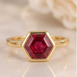 May include: A gold ring featuring a central hexagonal-cut deep red gemstone set in a smooth bezel setting. The band is polished yellow gold with a simple, clean design that highlights the geometric shape of the vibrant red stone at the centre.