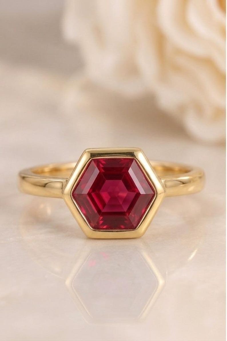 May include: A gold ring featuring a central hexagonal-cut deep red gemstone set in a smooth bezel setting. The band is polished yellow gold with a simple, clean design that highlights the geometric shape of the vibrant red stone at the centre.