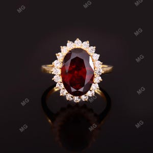 Puede incluir: Un anillo de oro con una gran piedra preciosa granate de color rojo intenso y corte ovalado en el centro. La piedra central está rodeada por un halo de pequeñas piedras blancas redondas, diamantes o circonitas, en una banda de oro amarillo.