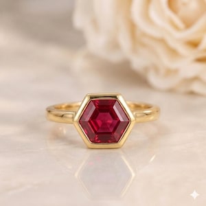 Peut inclure: Une bague en or avec une pierre précieuse rouge foncé taillée en hexagone au centre. L'anneau est lisse et poli, maintenant la pierre à six côtés dans un serti clos. Le design est symétrique, mettant en valeur la couleur rouge vive.