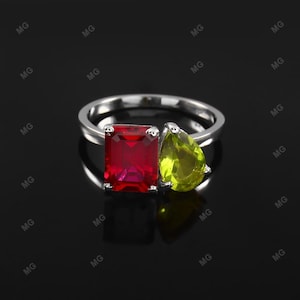 Op de afbeelding: Een zilveren ring met een rechthoekige rode edelsteen en een peervormige groene edelsteen. Het ontwerp van de ring is geplaatst tegen een donkere achtergrond, waardoor de kleuren van de edelstenen worden benadrukt.